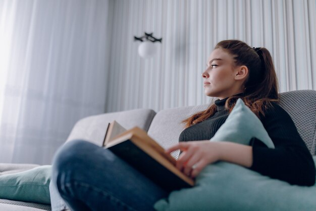 Aantrekkelijk meisje op de bank leest een papieren boek. geestelijke ontwikkeling. nuttig gebruik van tijd thuis. comfort voor thuis.