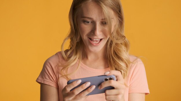 Aantrekkelijk opgewonden blond meisje emotioneel spelen in spel op smartphone over gele achtergrond