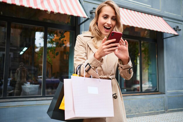 Aantrekkelijk opgewonden blond meisje in stijlvolle trenchcoat met boodschappentassen die vreugdevol mobiele telefoon gebruiken op straat in de stad