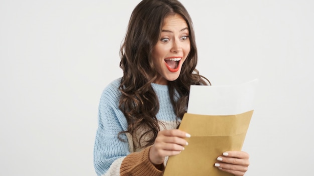 Aantrekkelijk opgewonden brunette meisje in trui die vreugdevol envelop opent met examenresultaten op camera geïsoleerd