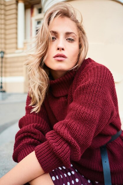 Aantrekkelijk stijlvol blond meisje in gebreide trui die in de camera kijkt terwijl ze sensueel buiten poseert