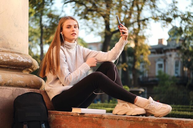 Aantrekkelijk studentenmeisje in een knusse trui die bedachtzaam in de camera kijkt terwijl ze selfie op de mobiele telefoon neemt tijdens de studiepauze buiten