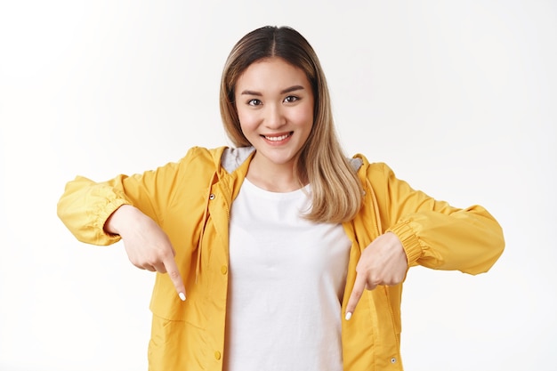 Aantrekkelijk vrolijk dwaas blond Aziatisch meisje dat naar beneden wijst wijsvinger kijk camera gelukkig optimistische glimlach stelt goede aanbeveling voor staande witte muur