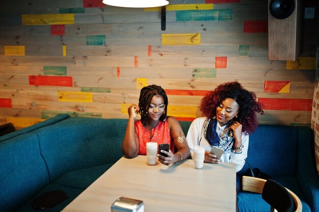 Aantrekkelijke Afro-Amerikaanse twee meisjesvrienden die in café zitten met latte en op mobiele telefoons kijken