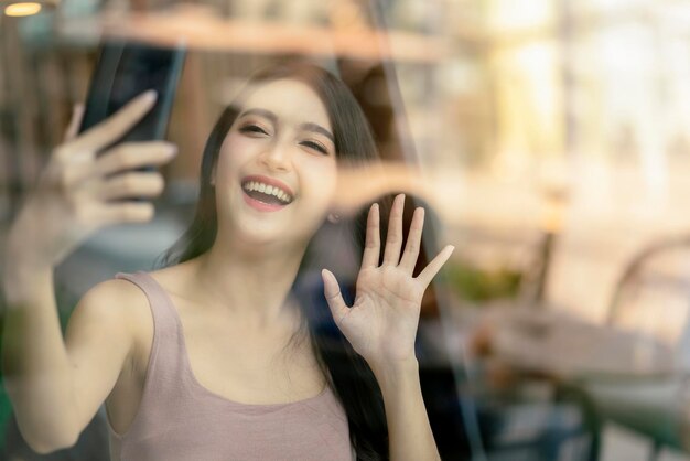 Aantrekkelijke aziatische vrouw die met de hand zwaait om hallo te zeggen tegen haar vriend via smartphone in café-restaurantfoto geschoten door raamwinkel