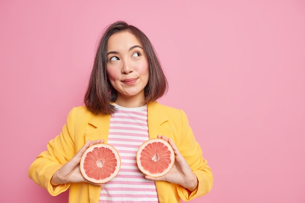Aantrekkelijke Aziatische vrouw heeft doordachte dromerige uitdrukking houdt grapefruit helften eet sappige citrusvruchten om calorieën te verbranden heeft gezonde voeding poses tegen roze muur met kopieerruimte