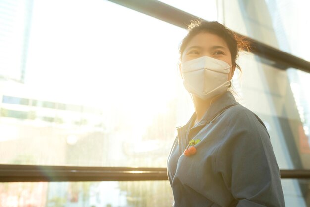 Aantrekkelijke Aziatische vrouwelijke vrouw casual doek dragen gezichtsmasker bescherming staande naast grote glazen moderne frame raam met zonlicht flare subset moment nieuwe normale levensstijl gezondheidszorg concept
