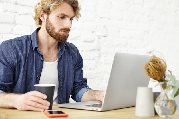 Aantrekkelijke bebaarde mannelijke ondernemer het drinken van thee of koffie tijdens het werken op generieke laptopcomputer tijdens de lunch op moderne café. ernstige jonge freelance werknemer met behulp van notebook pc voor verre werk