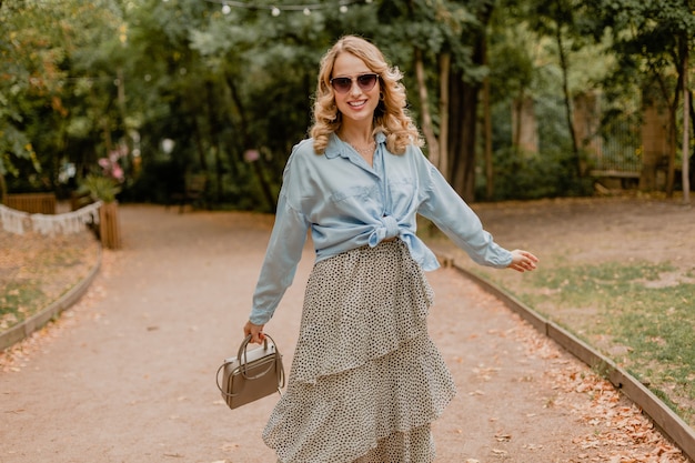 Aantrekkelijke blonde glimlachende vrouw die in park in de zomeroutfit loopt