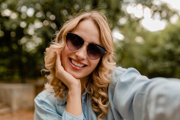 Aantrekkelijke blonde glimlachende vrouw die in park in zomeruitrusting loopt die selfiefoto op telefoon neemt