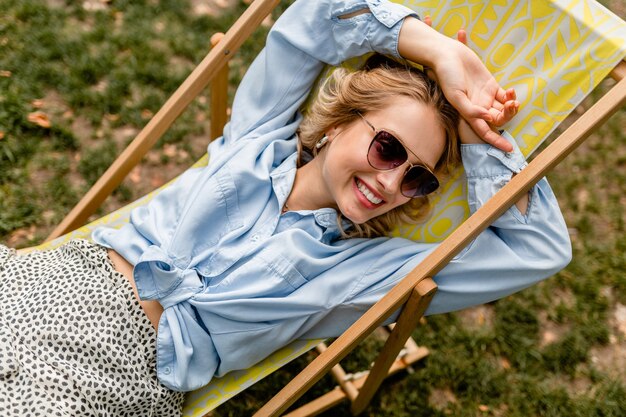 Aantrekkelijke blonde glimlachende vrouwenzitting in ligstoel in de zomeruitrusting