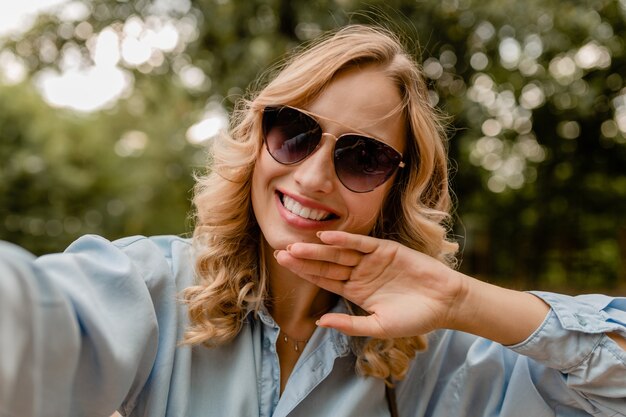 Aantrekkelijke blonde glimlachende witte tandenvrouw die in park in zomeruitrusting lopen die selfiefoto op telefoon neemt