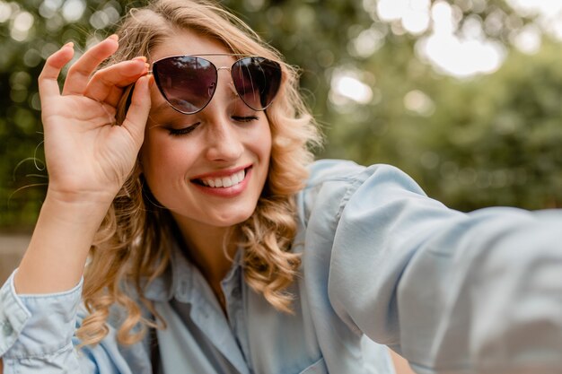 Aantrekkelijke blonde glimlachende witte tandenvrouw die in park in zomeruitrusting lopen die selfiefoto op telefoon neemt