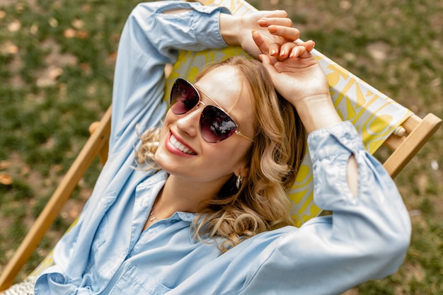 Aantrekkelijke blonde lachende vrouw zit ontspannen in een ligstoel in een stijlvolle outfit