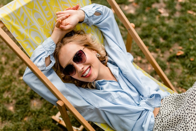 Gratis foto aantrekkelijke blonde lachende vrouw zit ontspannen in een ligstoel in een stijlvolle outfit