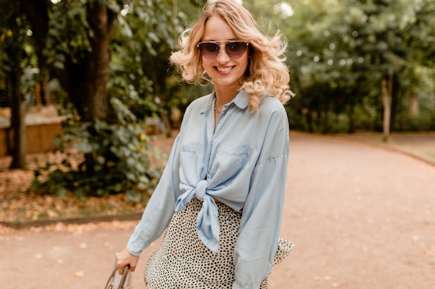 Aantrekkelijke blonde spontane vrouw wandelen in park in stijlvolle outfit elegante zonnebril en tas dragen