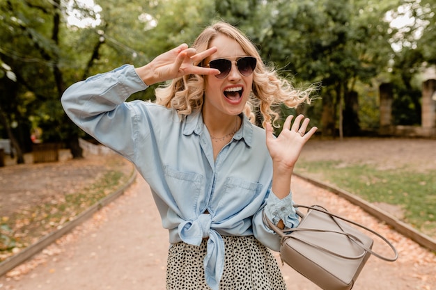 Aantrekkelijke blonde spontane vrouw wandelen in park in zomer outfit elegante zonnebril en tas dragen