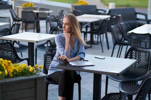 Aantrekkelijke blonde zakenvrouw denkt aan haar baan terwijl ze buiten in de cafetaria zit met papierwerk.