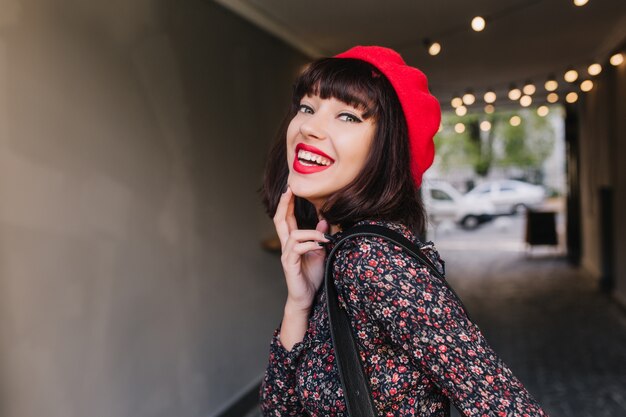 Aantrekkelijke brunette meisje in stijlvolle vintage outfit poseert koket met oprechte glimlach haar kin aan te raken. Close-up portret van elegante jonge vrouw in trendy Franse kleding lachen en plezier maken