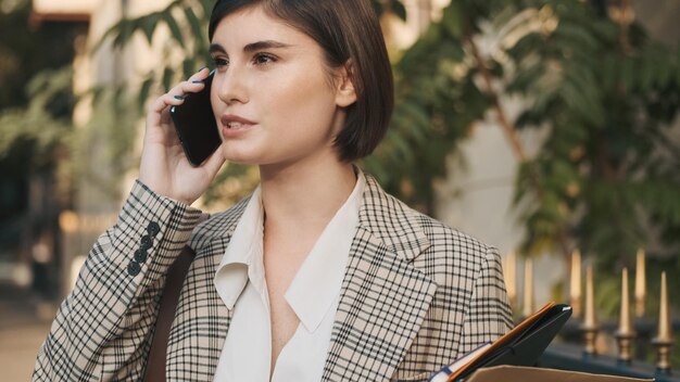 Aantrekkelijke brunette zakenvrouw die er stijlvol uitziet en een zakelijk gesprek voert op straat in de stad Jonge dame die buiten aan de telefoon praat