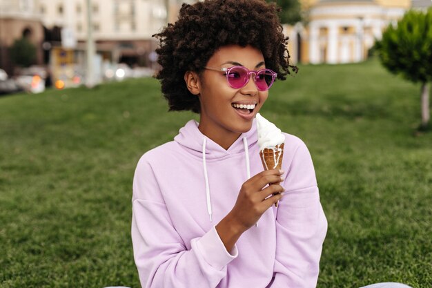 Aantrekkelijke charmante gelukkige vrouw in roze zonnebril, stijlvolle oversized hoodie lacht en eet buiten ijs