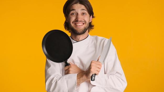 Gratis foto aantrekkelijke chef-kok die een uniform mes en een koekenpan draagt en de handen over de kleurrijke achtergrond houdt jonge glimlachende man klaar om te koken
