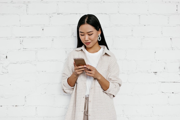 Aantrekkelijke dame in beige vest en t-shirt chats in telefoon op witte bakstenen muur muur