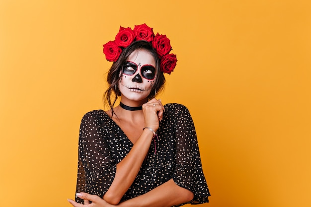 Aantrekkelijke dame met Halloween-make-up zorgvuldig opgezocht. Portret van meisje met rode bloemen in haar haar die zich voordeed op oranje achtergrond.