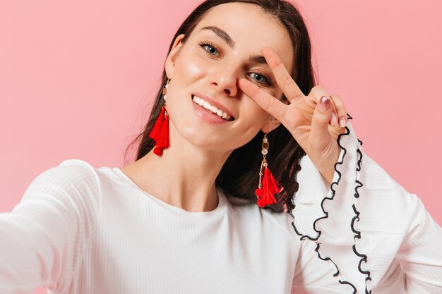 Aantrekkelijke dame met rode grote oorbellen glimlacht en maakt selfie op roze achtergrond.