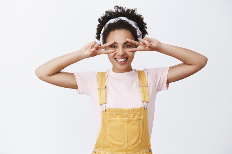 Gratis Foto aantrekkelijke donkere stijlvolle vrouwelijke vrouw in hoofdband over haar en gele trendy overall, vredesgebaar over ogen tonen en glimlachend met zorgeloze en gelukkige uitdrukking