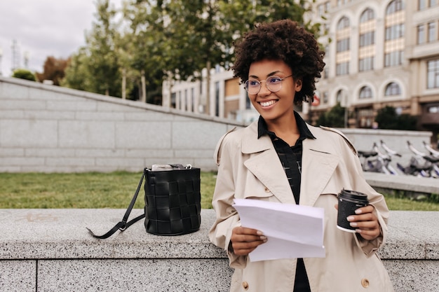 Aantrekkelijke donkere vrouw in beige trenchcoat en bril glimlacht, houdt koffiekopje en vel papier buiten vast