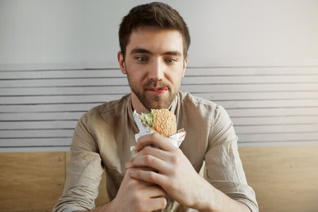 Aantrekkelijke donkerharige man zitten in cafe, kijken met een gelukkige uitdrukking op sandwich, gelukkig om iets te eten na de hele dag op het werk. Hongerige mens die hamburger gaat eten.