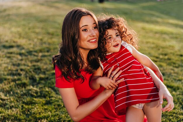 Aantrekkelijke donkerharige vrouw zittend op het gras met dochter. Buiten schot van lachen krullend meisje zusje omarmen op aard