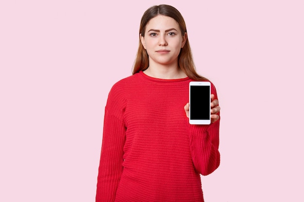 Aantrekkelijke ernstige jonge Europese tiener met lang haar, gekleed in rode losse trui, houdt moderne mobiele telefoon met zwart leeg