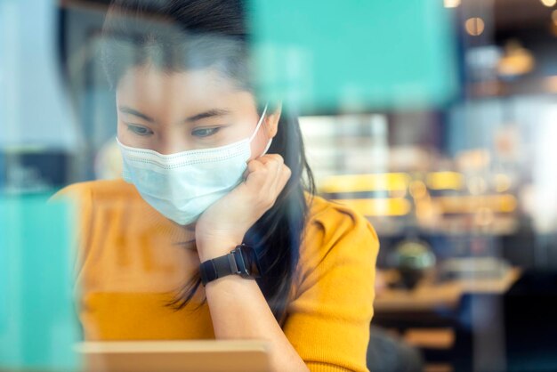 Aantrekkelijke freelance aziatische vrouwelijke creatieve persoon draagt een casual gele doek en gezichtsmasker bescherming sociale afstand in gedeelde ruimte coworking ruimte nieuwe levensstijl met doordachte en frisheid emotie