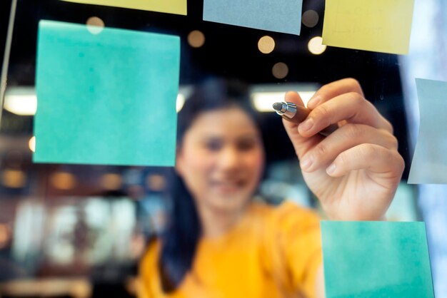 Aantrekkelijke freelance aziatische vrouwelijke creatieve persoon draagt een casual gele stoffen handpunt en noteert een papieren notitiestokherinnering bij een nieuwe levensstijl in een coworking-ruimte met een doordachte en frisheidemotie