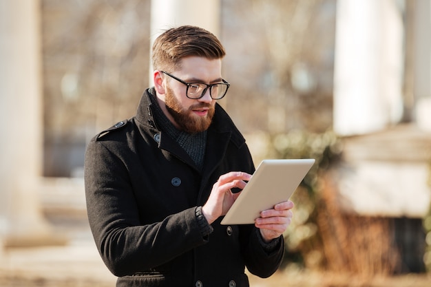 Gratis foto aantrekkelijke gebaarde jonge mens die tabletcomputer in openlucht met behulp van