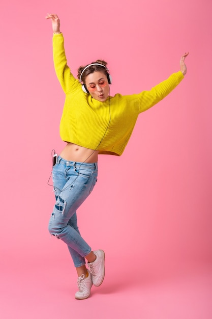 Aantrekkelijke gelukkig grappige vrouw dansen luisteren naar muziek in koptelefoon gekleed in hipster kleurrijke stijl outfit geïsoleerd op roze muur, gele trui en zonnebril dragen, plezier