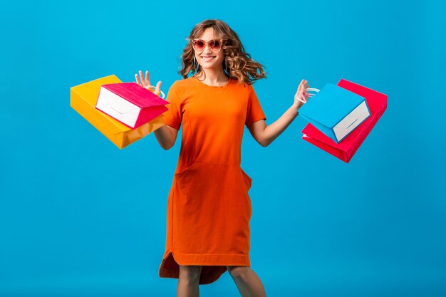 Aantrekkelijke gelukkig lachend stijlvolle vrouw shopaholic in oranje trendy oversized jurk springen met boodschappentassen bedrijf op blauwe achtergrond geïsoleerd