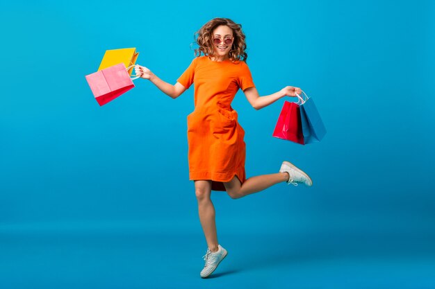 Aantrekkelijke gelukkig lachend stijlvolle vrouw shopaholic in oranje trendy oversized jurk springen met boodschappentassen op blauwe studio achtergrond geïsoleerd