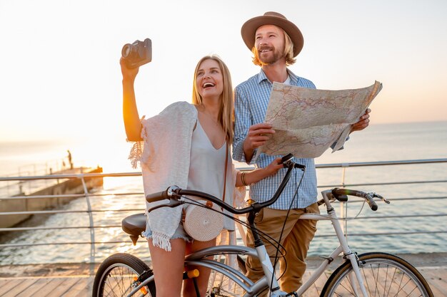 Aantrekkelijke gelukkige paar reizen in de zomer op de fiets, man en vrouw met blond haar boho hipster stijl mode samen plezier, op zoek in kaart sightseeing fotograferen op camera