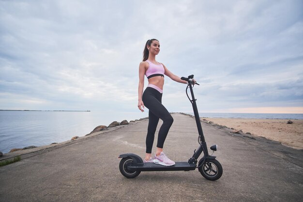 Aantrekkelijke gelukkige vrouw geniet van het ochtendrijden met haar nieuwe elektrische scooter aan zee.