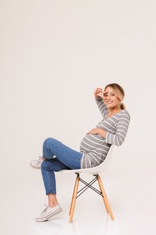 Aantrekkelijke glimlachende jonge zwangere vrouw zittend op een stoel geïsoleerd, vredesgebaar tonend