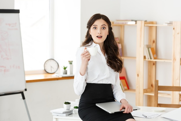 Aantrekkelijke glimlachende zakenvrouw die aan het bureau zit en een kopje koffie vasthoudt, ze ontspant en