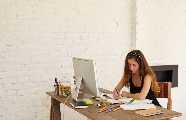 Aantrekkelijke grafisch ontwerper vrouw trekt schetsen van nieuw logo voor tandheelkundige kliniek zit aan het bureau met pc-computer, documenten en gekleurd briefpapier. Kopieer de ruimtemuur voor reclame-inhoud of tekst