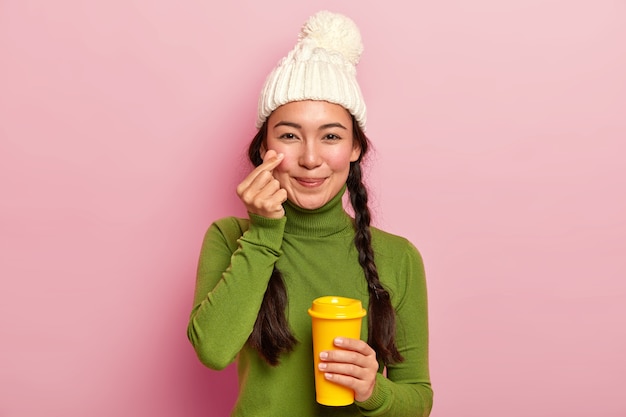 Aantrekkelijke jonge Aziatische meisje maakt Koreaans als teken, vormt klein hartje in de buurt van Wang, spreekt liefde uit aan vriendje, draagt warme muts en casual trui, drinkt afhaalmaaltijden koffie