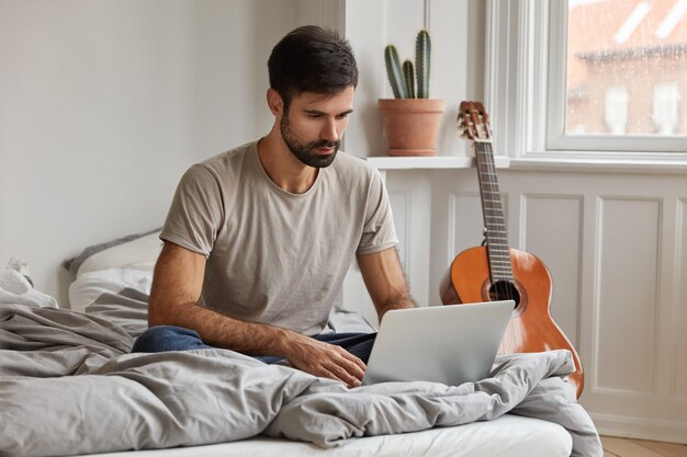 Aantrekkelijke jonge bebaarde freelancer zit voor geopende laptopcomputer, houdt benen gevouwen in comfortabel bed