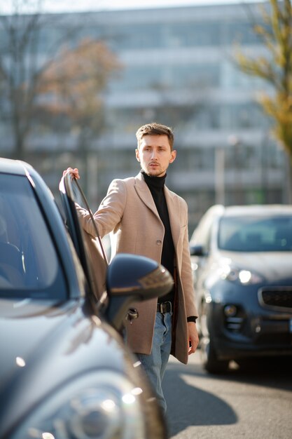 Aantrekkelijke jonge blanke man opent een autodeur, gekleed in beige jas in het centrum op een zonnige dag