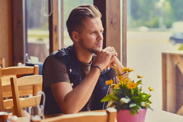 Aantrekkelijke jonge blonde bebaarde man gekleed in een leren jas zit aan de tafel in een café.