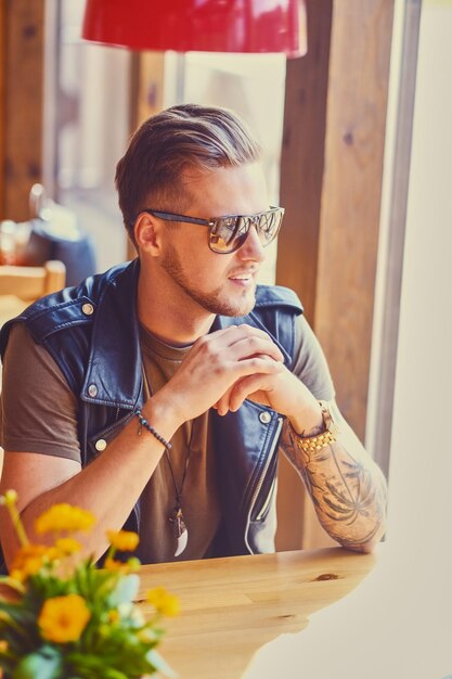 Aantrekkelijke jonge blonde bebaarde man gekleed in een leren jas zit aan de tafel in een café.